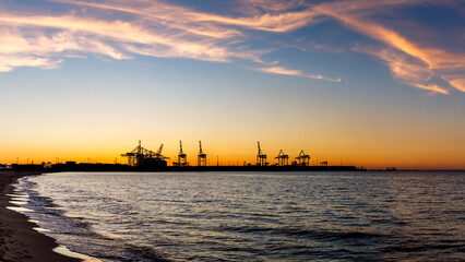 Sunset over harbor in Gdansk with commercial dock and cranes. View from Stogi beach in Baltic sea. Silhouette of industrial port - obrazy, fototapety, plakaty