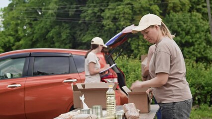 Volunteers prepare and pack humanitarian aid, long-lasting food in cardboard boxes and put them in the trunk of the car. Group of food bank volunteers help to sort tinned food for people in poverty - Powered by Adobe