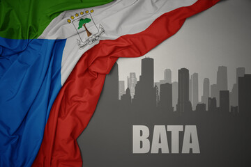 abstract silhouette of the city with text Bata near waving colorful national flag of equatorial guinea on a gray background.