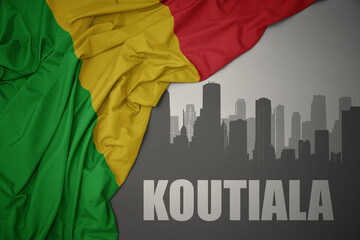 abstract silhouette of the city with text Koutiala near waving colorful national flag of mali on a gray background.