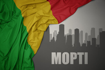 abstract silhouette of the city with text Mopti near waving colorful national flag of mali on a gray background.
