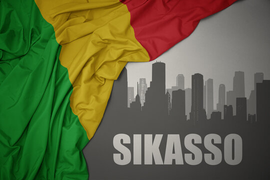 abstract silhouette of the city with text Sikasso near waving colorful national flag of mali on a gray background.