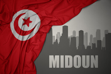 abstract silhouette of the city with text Midoun near waving colorful national flag of tunisia on a gray background.