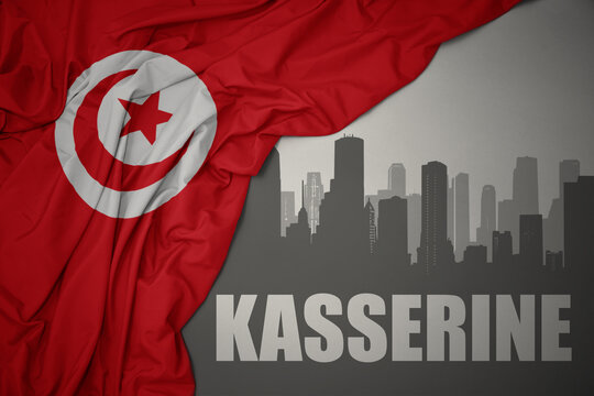 abstract silhouette of the city with text Kasserine near waving colorful national flag of tunisia on a gray background.