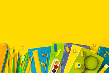 The concept of back to school. Colorful school supplies in blue, green on a yellow background. Books, notebooks, pens, paper clips, paints and magnifying glass. Top view, flat lay, copy space