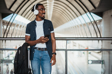 Happy african man listening to music on wireless headphones while he is waiting at a train station