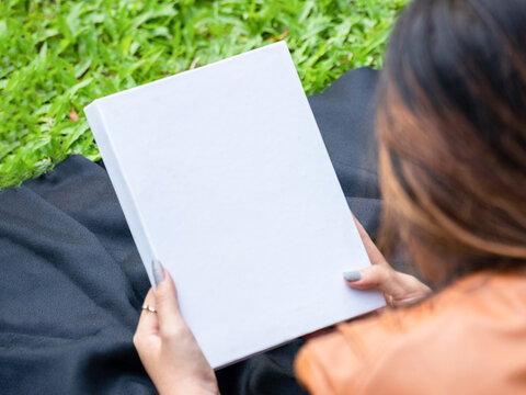 Mockup of a white blank hardcover book featuring a woman reading it at a park