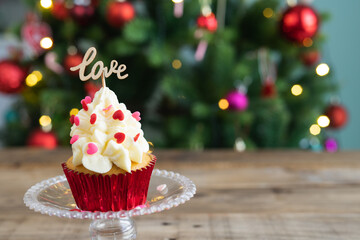 Cupcake in display stand on wooden background with LOVE sign and out-of-focus Christmas tree in the background. Copy space.