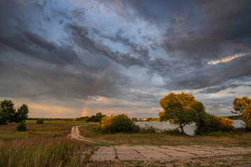 Rainbow against a dramatic sky. Picturesque rural landscape. The trees in the foreground are illuminated by the rays of the setting sun.