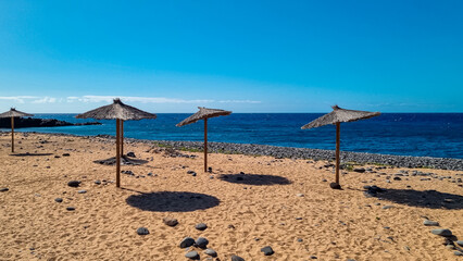 Straw sun umbrellas on sand beach connected to stone pebble beach Playa de San Blas near coastal village Los Abrigos, Tenerife, Canary Islands, Spain, Europe, EU. Coastline of Atlantic Ocean. Vacay