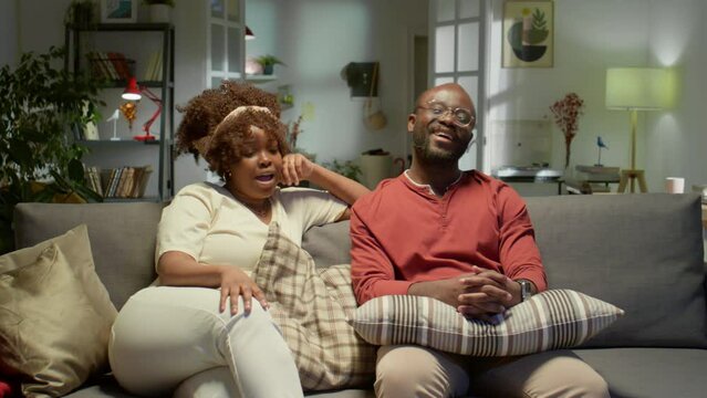 Happy African American couple resting on sofa at home, smiling and chatting while watching TV together