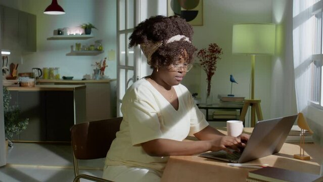 Zoom in shot of young African American woman in glasses typing on laptop at desk and drinking tea while working online in evening at home
