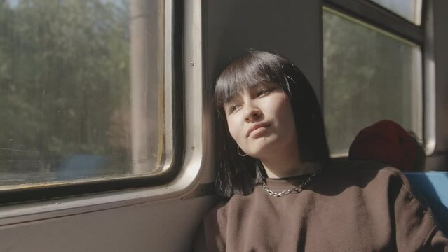 A young woman rides in an electric train, sits with her head tilted to the window, looks out the window, closes her eyes. Outside the window is a forest,the sun is shining. Slow motion 4k footage