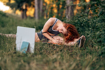 Young caucasian woman doing fitness stretching yoga exercise in the forest while using tablet for online class or virtual tutorials. healthy lifestyle.