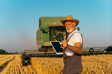 Senior farmer in wheat field holding digital tablet and showing thumb up. He is smiling. Combine...