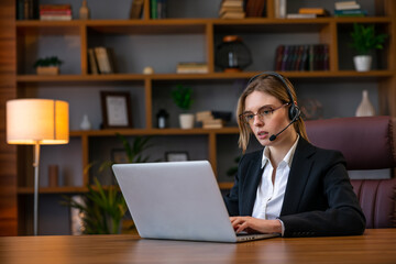 Fototapeta premium Attractive freelancer female talking in a video conference on line with a headset with microphone and laptop in an office desktop or home desk