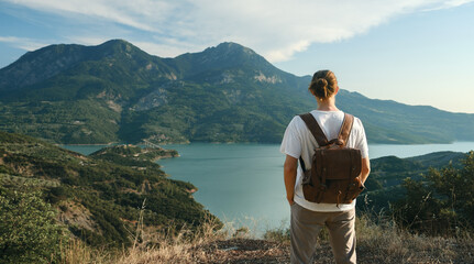 Back view of wanderlust man with backpack looking at scenic view of lake and mountains from a view point. - obrazy, fototapety, plakaty