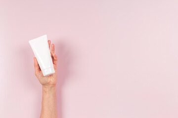Woman hand holding white blank plastic cosmetics cream tube on pastel pink background. Skincare,...