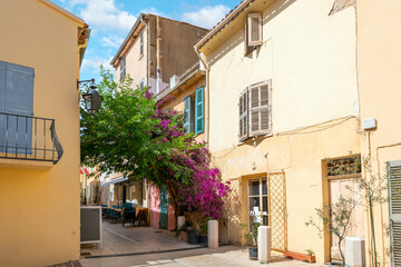 A picturesque alley with purple Bougainvillea flowers covering the entry and a sidewalk cafe with tables in the old town village section of Saint-Tropez, France, on the French Riviera.