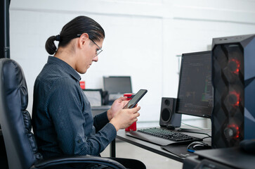 Un hombre joven viendo su celular sentado frente a una computadora
