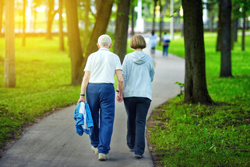 Fototapeta premium Elderly couple holding hands while walking together outdoors. Senior couple walking outdoors at park at summer sunny day, active lifestyle. Senior couple holding hands while walking on fresh air.