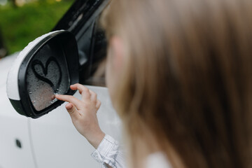 A young girl draws a heart on the side window of a car in the summer. Love concept.