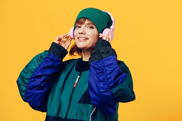 a happy, joyful woman in a green jacket and hat enjoys music standing in pink headphones on a yellow background, holding them with her hands and looking at the camera. Horizontal photo