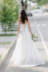 Fototapeta na wymiar A stylish European bride in a white dress with a white bouquet is standing on the street. Photo from the back