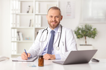 Smiling young male doctor sitting in an office and looking at camera and writing