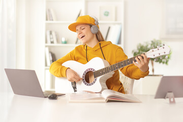 Young female with an acoustic guitar and headphones singing in front of a laptop computer