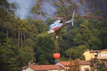 Un elicottero sta portando un bucket con acqua per spengere il fuoco tra il bosco e le case, Toscana, Italia