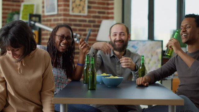 Diverse group of people having fun together at home, laughing and enjoying alcoholic drinks. Cheerful friends drinking beer and eating chips, smiling and playing with food for entertainment.
