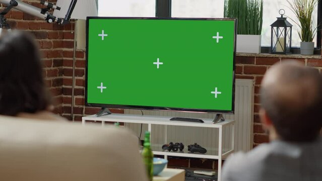 Young people on sofa looking at television with green screen display, watching isolated chroma key template with mockup background. Friends using blank copy space on TV. Close up.