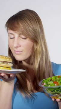PORTRAIT, CLOSE UP: Thoughtful young woman chooses a salad over a burger while weighing her lunch options. Beautiful Caucasian female opts to eat a healthy green salad over a fatty double hamburger.