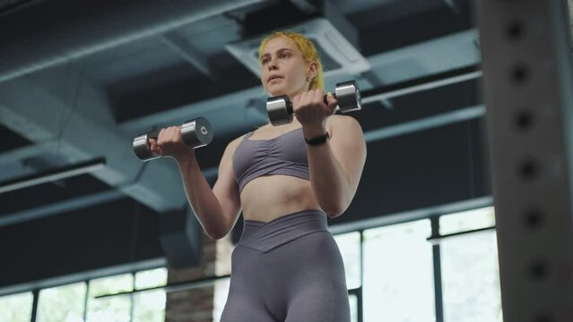 Young athletic woman with green hair lifting dumbbells during muscle workout for arms in gym