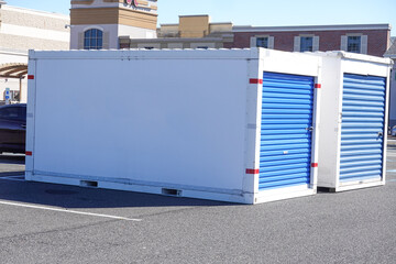 Two white storage pods with  blue doors on a asphalt road in front of some stores - obrazy, fototapety, plakaty