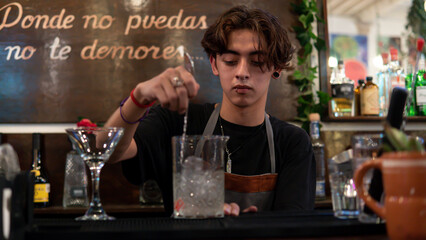 Bartender mixing a drink with ice at the bar counter. There are plants behind and a text.