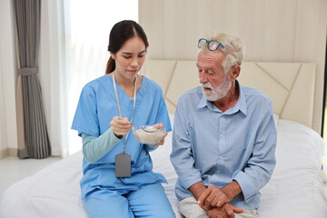 Happy caregiver feeding senior old man having cancer on bed with rice porridge soup for breakfast...