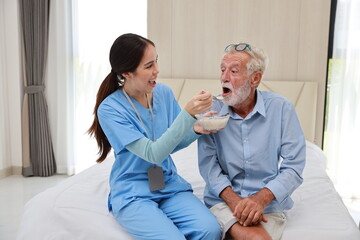 Obraz premium Happy caregiver feeding senior old man having cancer on bed with rice porridge soup for breakfast at retirement house. Asian smiling nurse taking good help care and support of elder patient at home.
