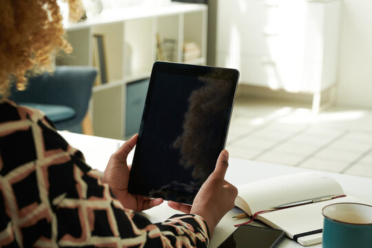 Close-up of African girl looking at screen of digital tablet and working online sitting at table with notepad in the room