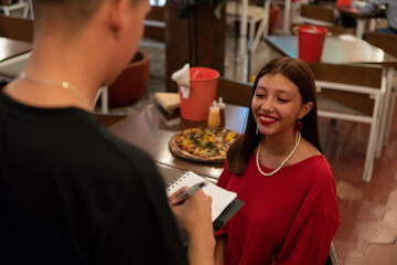 Smiling young Latin woman in a dress and red lipstick is sitting at a restaurant table asking the waiter for an order.