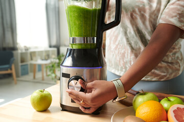 Close-up of young girl turning on the button on blender to make green vitamin smoothie at table...