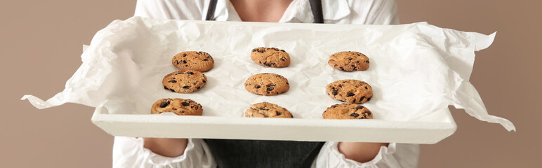 Female baker holding tray with tasty cookies on brown background, closeup