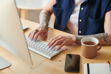 Close-up of young woman with tattoos on her hands typing on keyboard at table and drinking tea, she doing her online work on computer