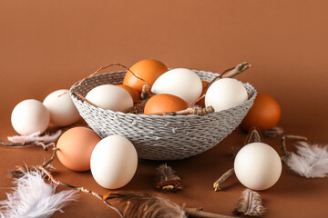 Wicker bowl with chicken eggs, feathers and tree branches on color background