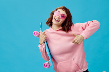 Funny joyful pretty redhead lady in pink hoodie sunglasses with penny board smiling posing isolated on blue studio background. Copy space Banner Offer. Fashion Cinema. Holiday activity