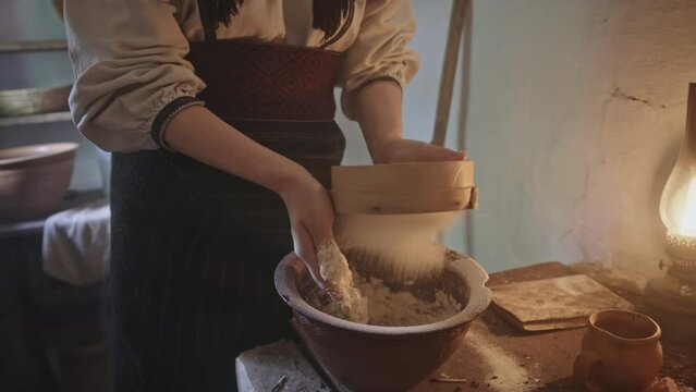 Slavic woman sieving flour into dough. Tilt up and down view of female in traditional Ukrainian clothes and headscarf sieving dough over bowl and kneading dough while preparing pastry in small kitchen