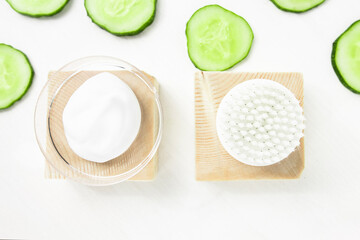 Body cream sample and cleansing brush with pieces of raw fresh green cucumber on a white wooden table. Moisturizing, nourishing body treatment.