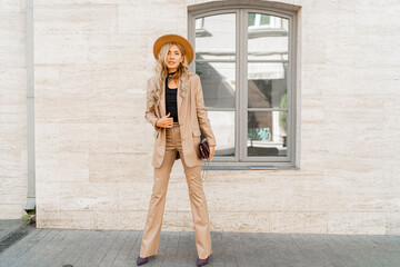 Fashion outdoor photo of stylish blond woman with perfect wavy hairs in beige hat and casual suit. Full lenght.