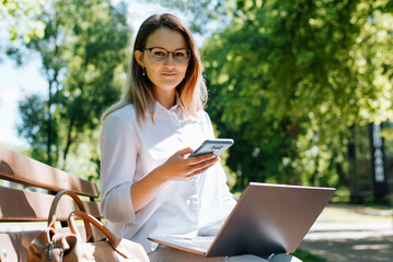 Obraz premium Stylish pretty female student freelancer in eyeglasses use laptop, hold mobile phone while relaxing on bench in park and looking at camera. Smiling young woman working remotely on netbook, e-learning
